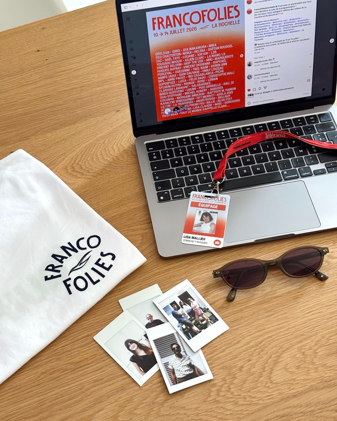 Sur un bureau en bois : un ordinateur ouvert sur l’affiche des Francofolies 2026, un badge, un tee-shirt Francofolies, des lunettes de soleil et des photos instantanées.