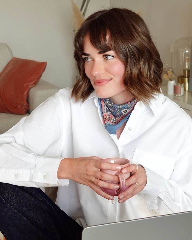 Lisa en chemise blanche et foulard, une tasse entre les mains, le regard tourné vers la fenêtre.