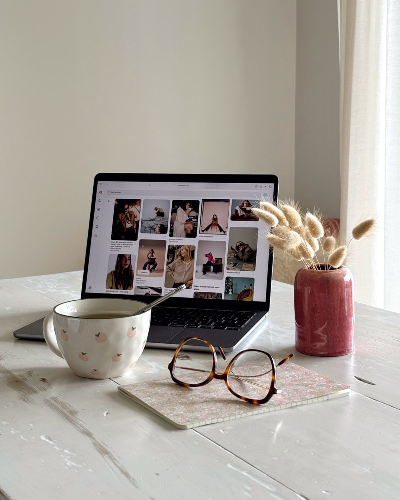 Un ordinateur portable ouvert sur une grille de contenus, une tasse, des lunettes et un bouquet de fleurs séchées.