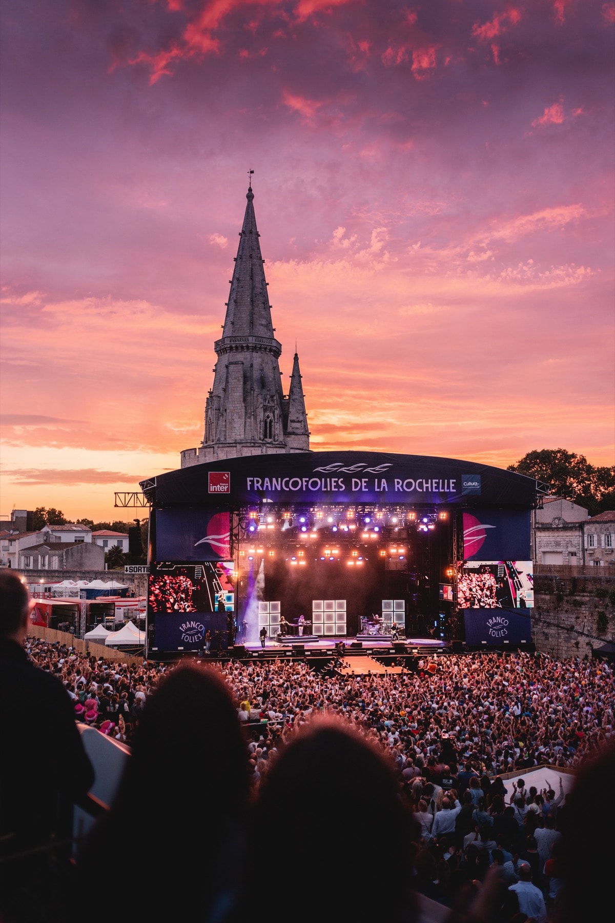 La grande scène des Francofolies au coucher de soleil, la flèche de l’église Saint-Sauveur derrière, et la foule bras levés.
